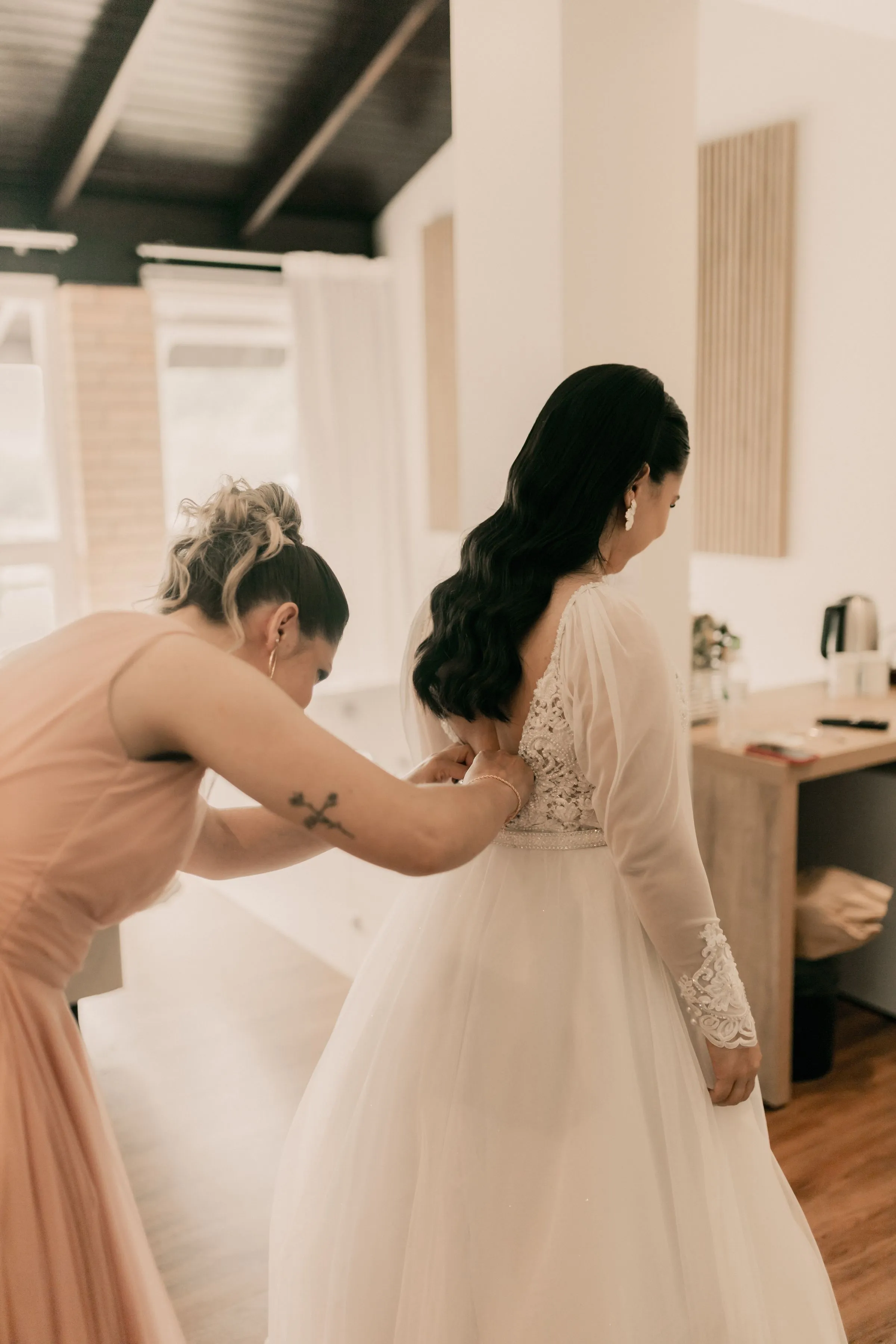 A bridesmaid in blush pink laces the back of the bride’s dress in a bright bedroom