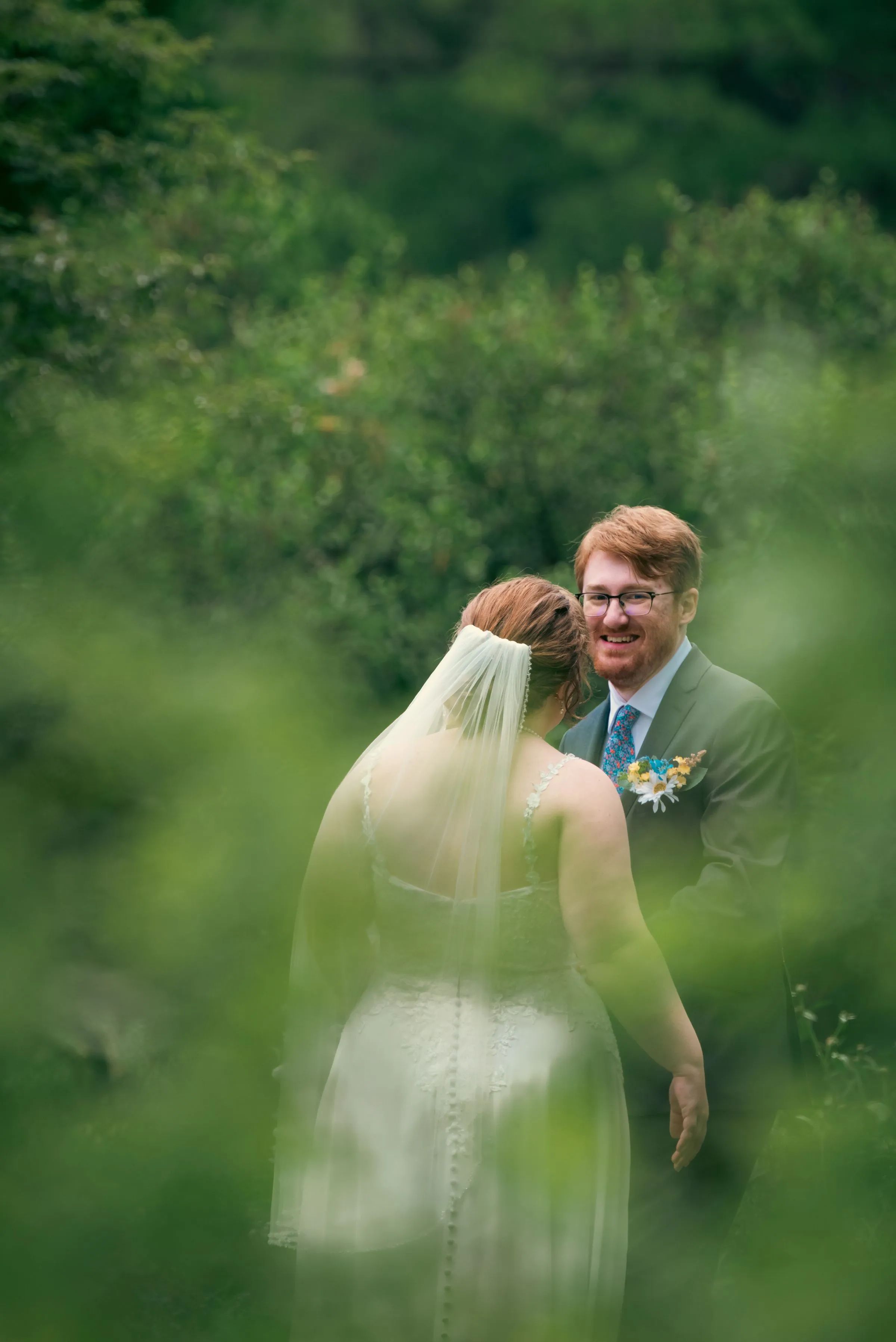 The couple talk quietly among tall green plants, seen through the leaves
