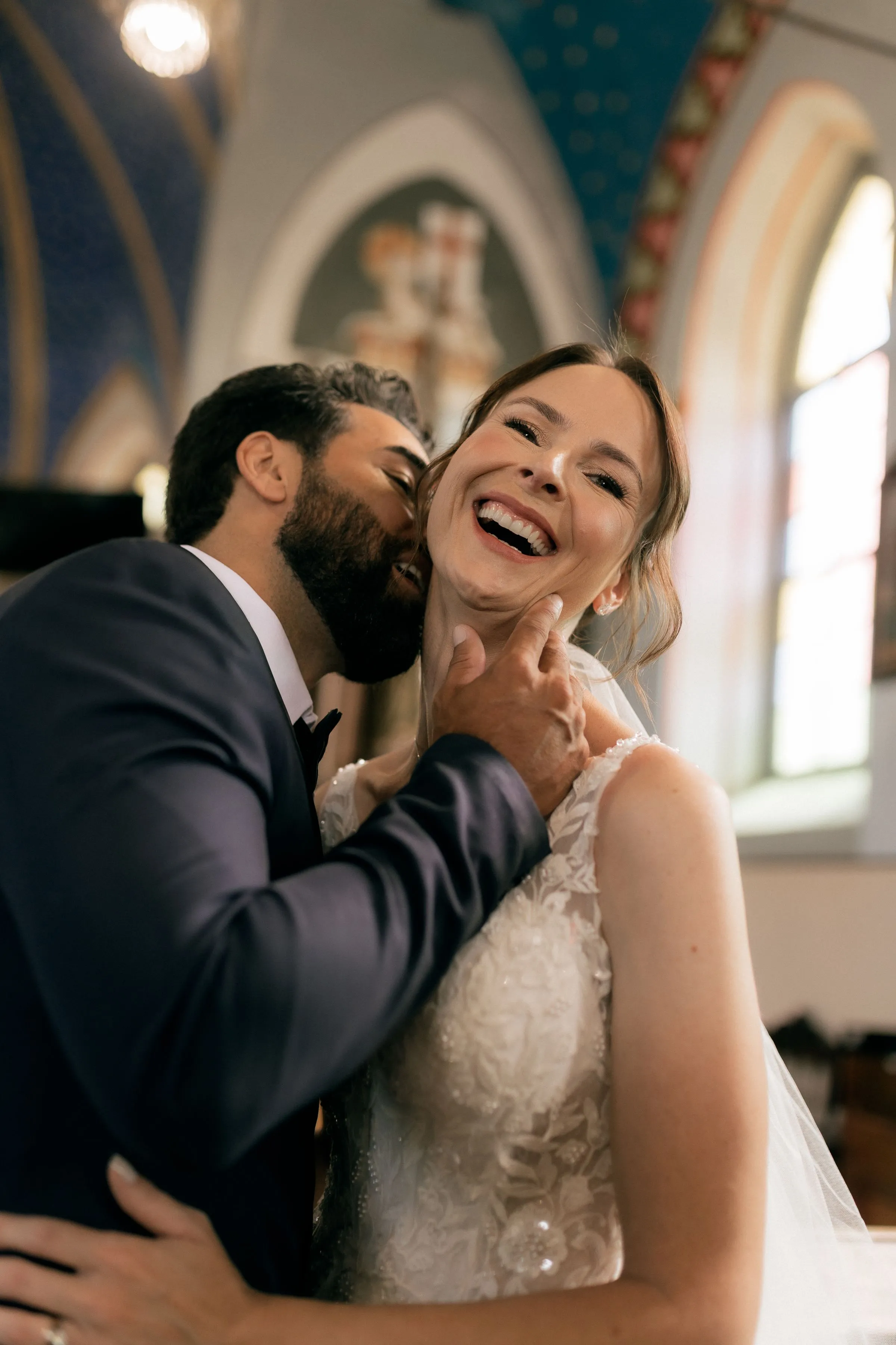 The groom kisses the bride’s cheek as she laughs, in a church with arched windows