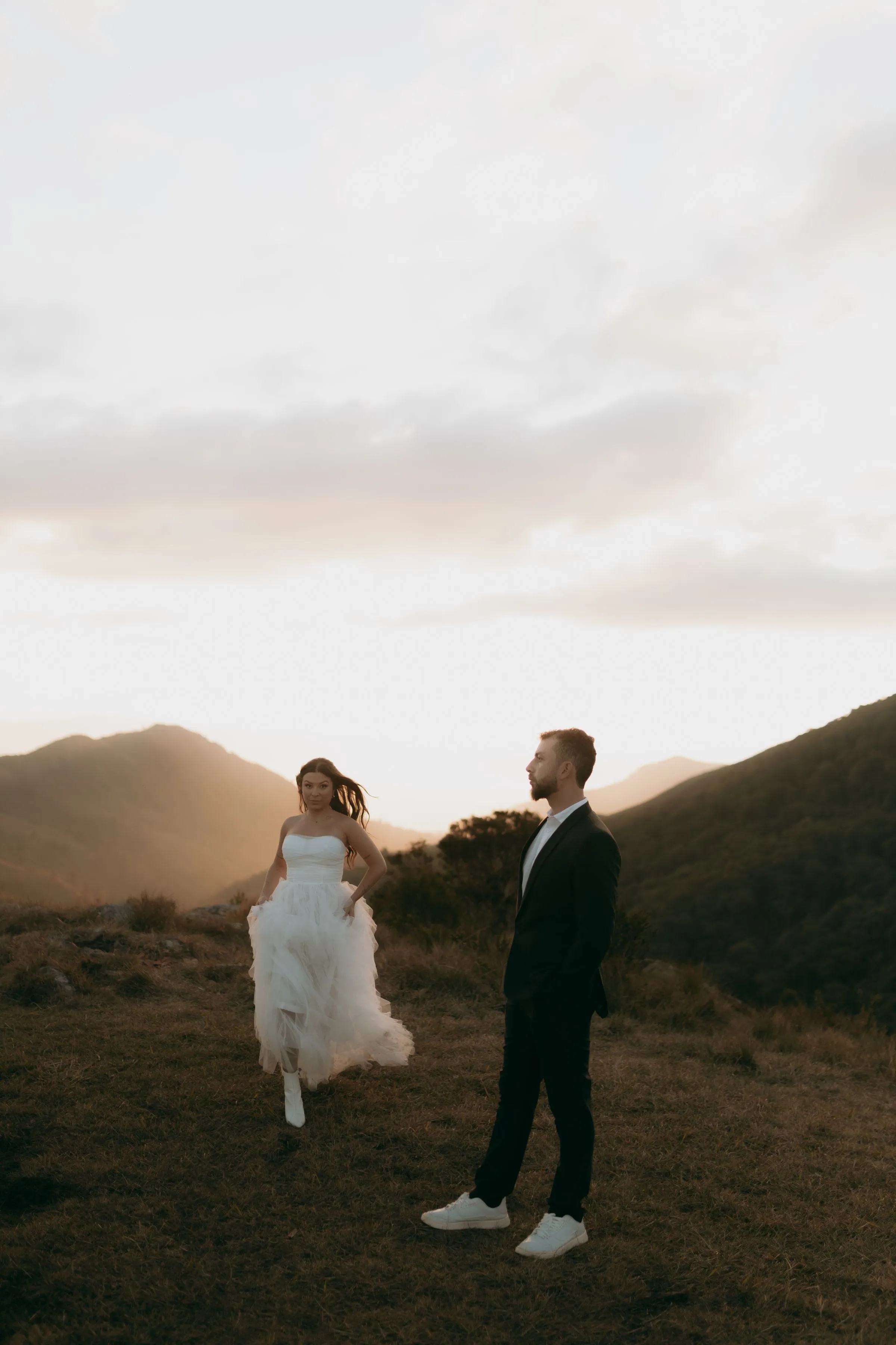The bride runs barefoot across a grassy ridge towards the groom at sunset