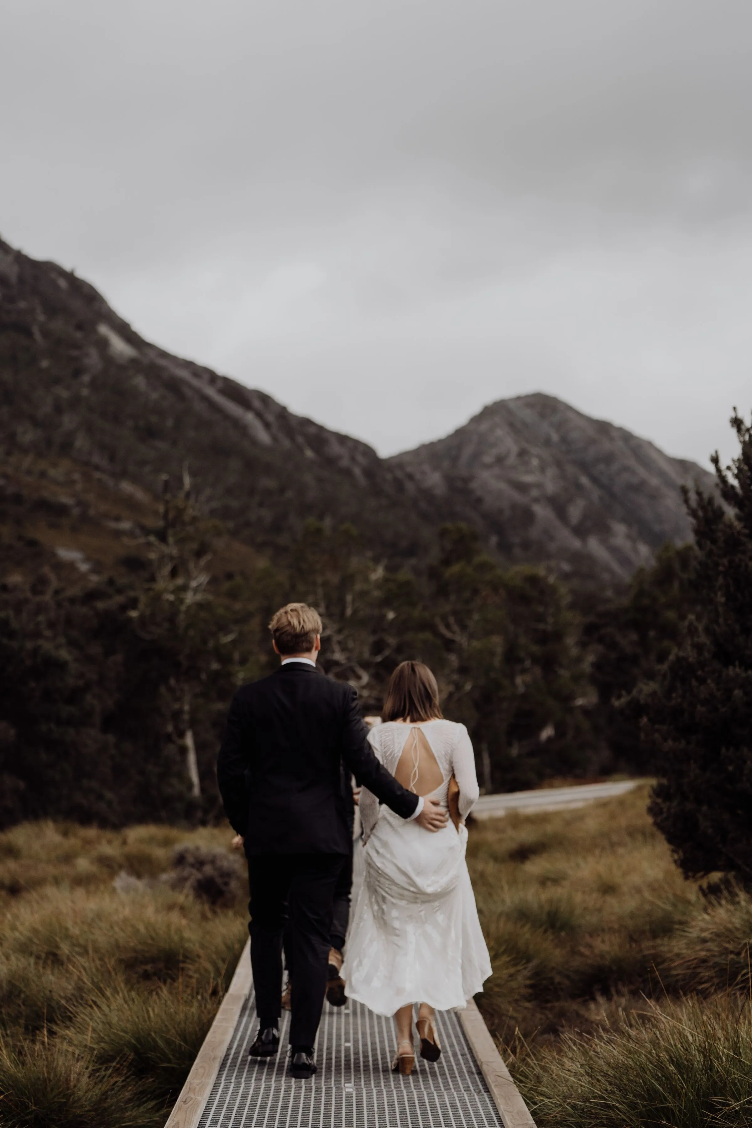 A couple in wedding clothes walk along a boardwalk towards the mountains