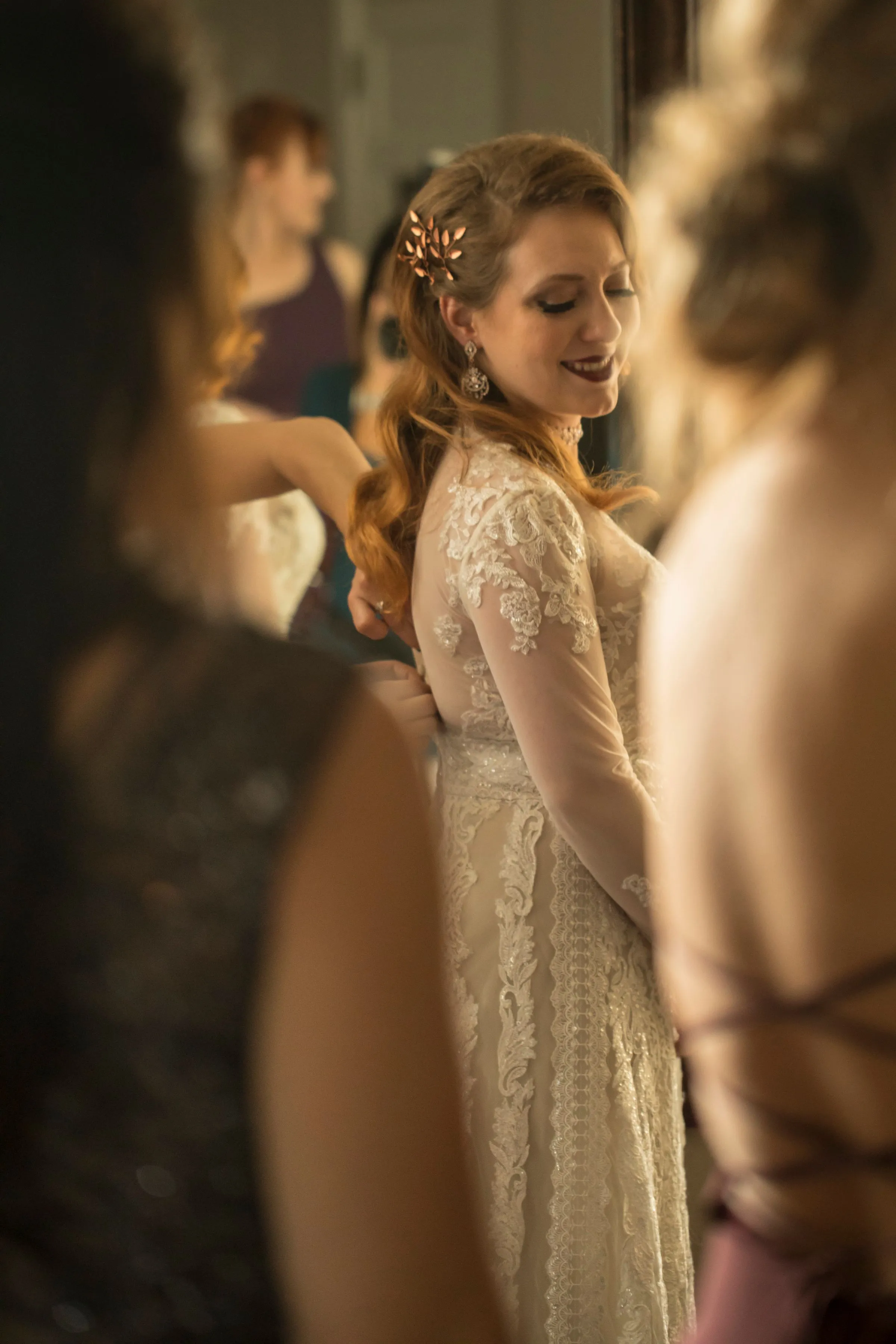 The bride, in a lace dress, looks down and smiles as friends help her get ready in warm light