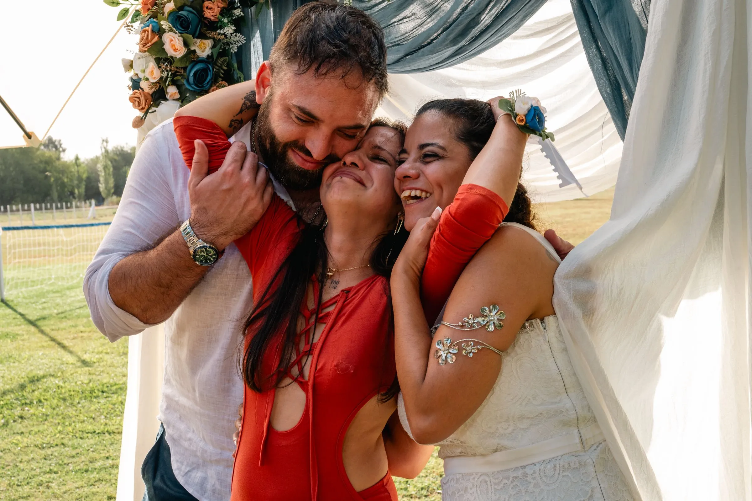 The couple hug a friend under a floral arch, everyone laughing