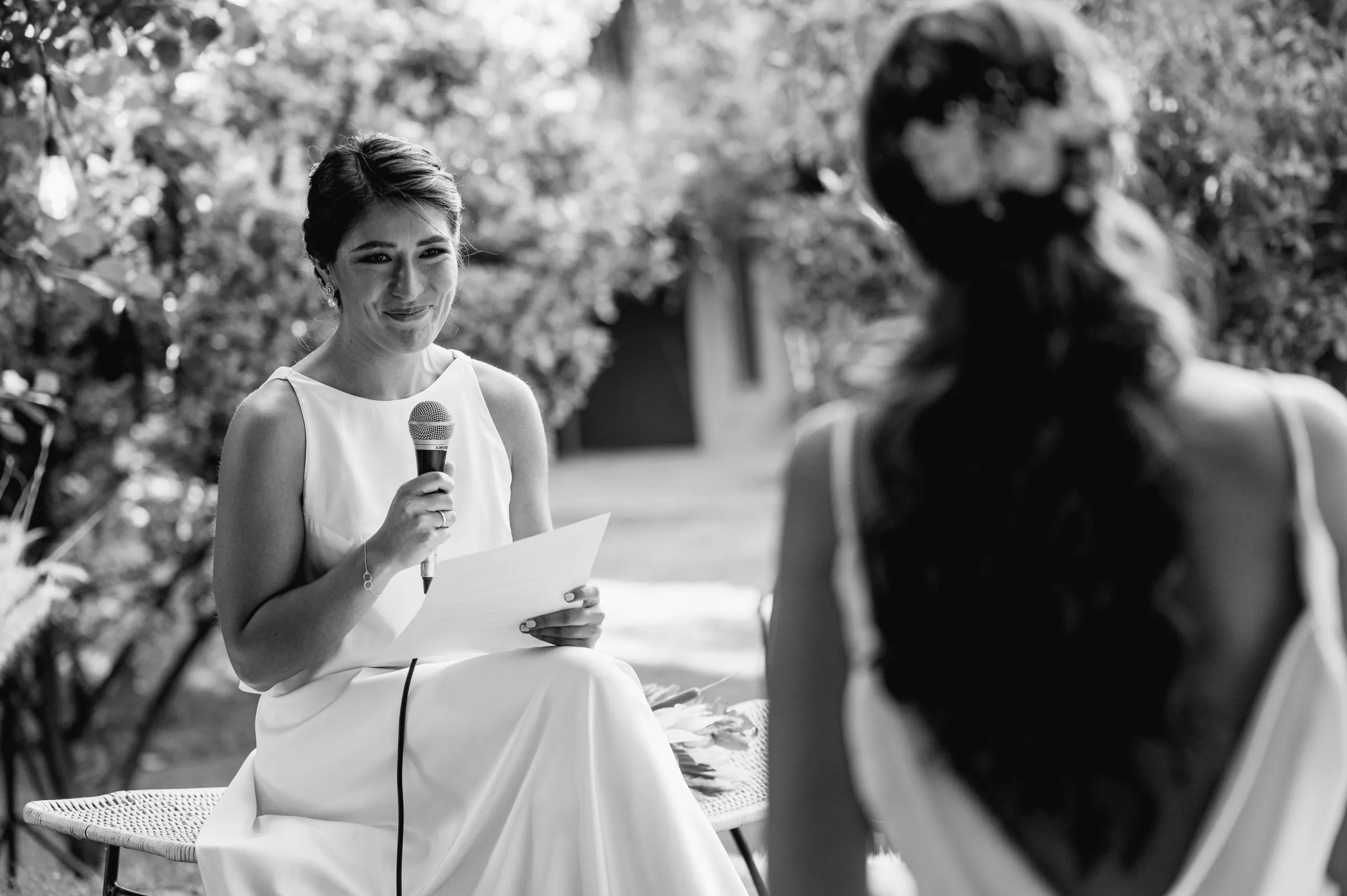 Black and white photo of the maid of honour reading her speech, the bride watching from behind