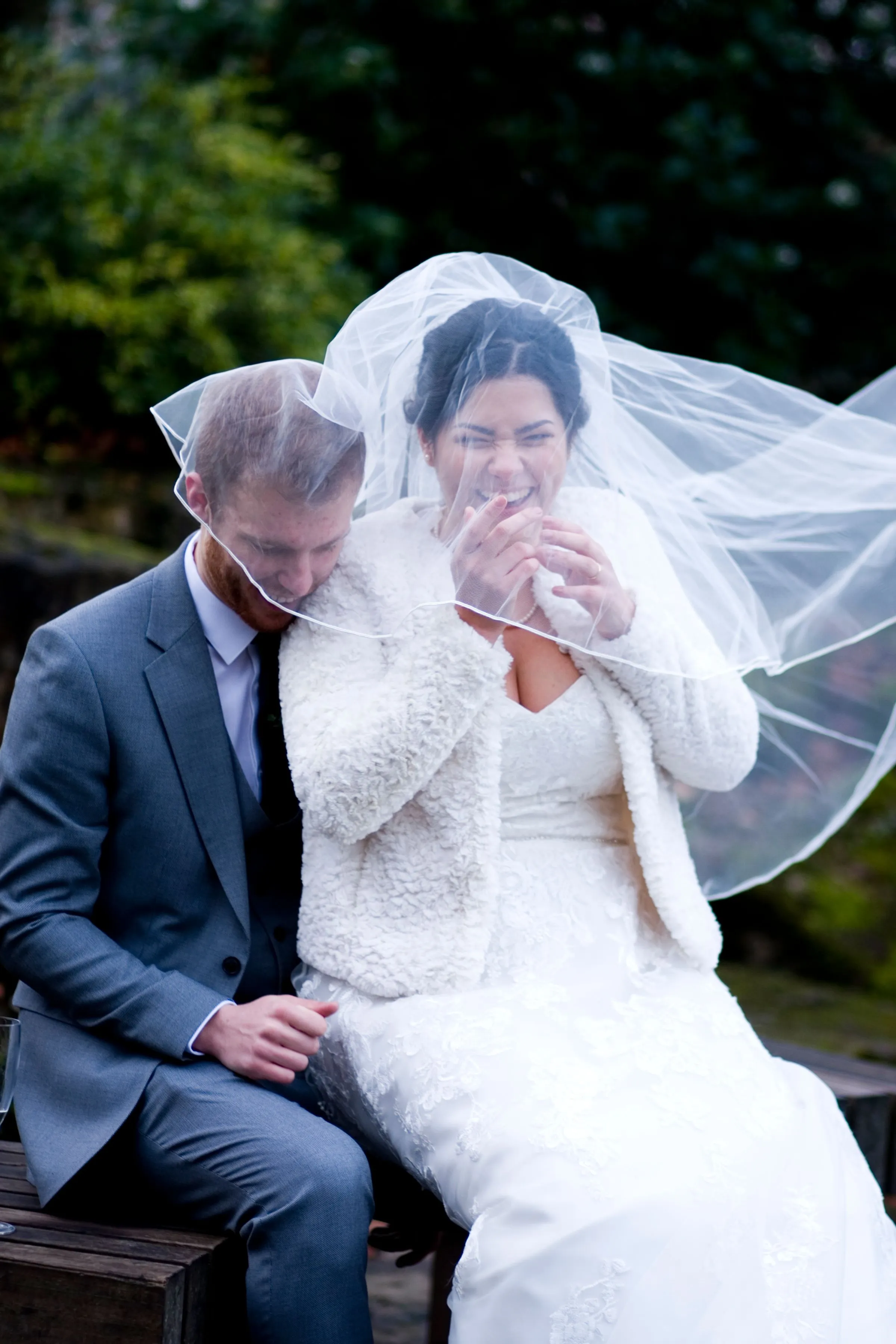 The bride laughs as the wind blows her veil over the groom, the two of them sitting on a bench under trees